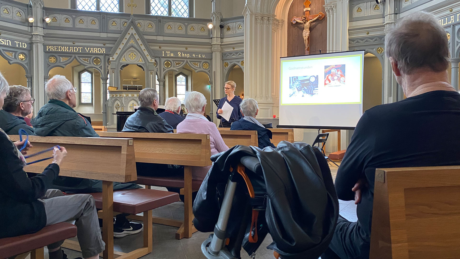 Bild på när katarina strandberg håller i 60-talstemat som var en av hemtjänstens kundaktiviteter, denna gång i ersta kyrka. Man ser publiken som sitter i kyrkbänkarna, med en rullator i förgrunden.
