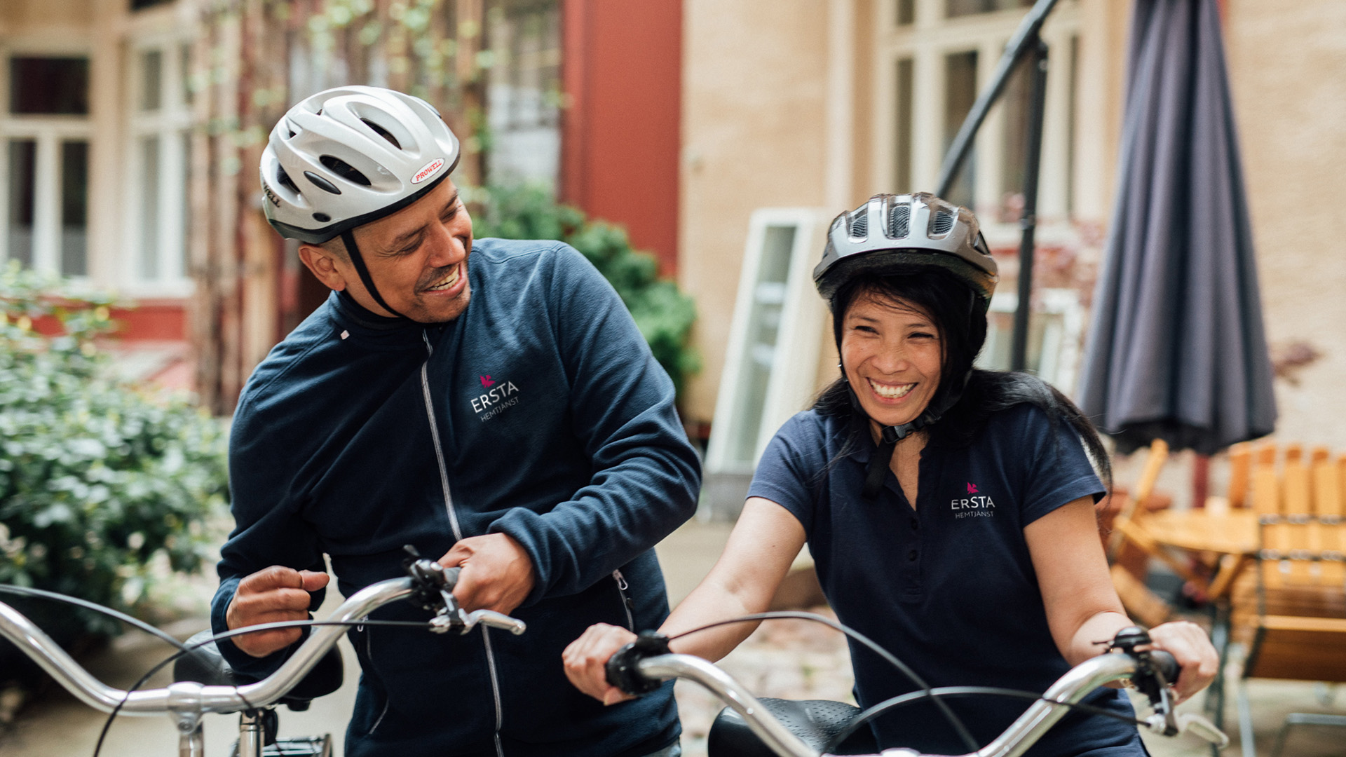 Två glada hemtjänstmedarbetare med cykelhjälmar håller varsin cykel