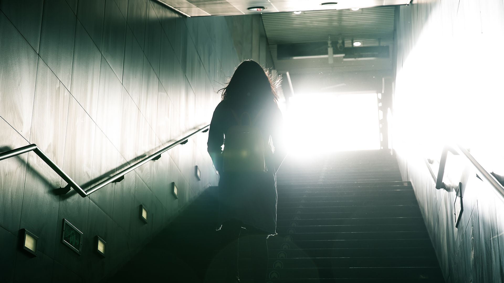 Kvinna i motljus på väg upp för en trappa, från mörker mot ljus.